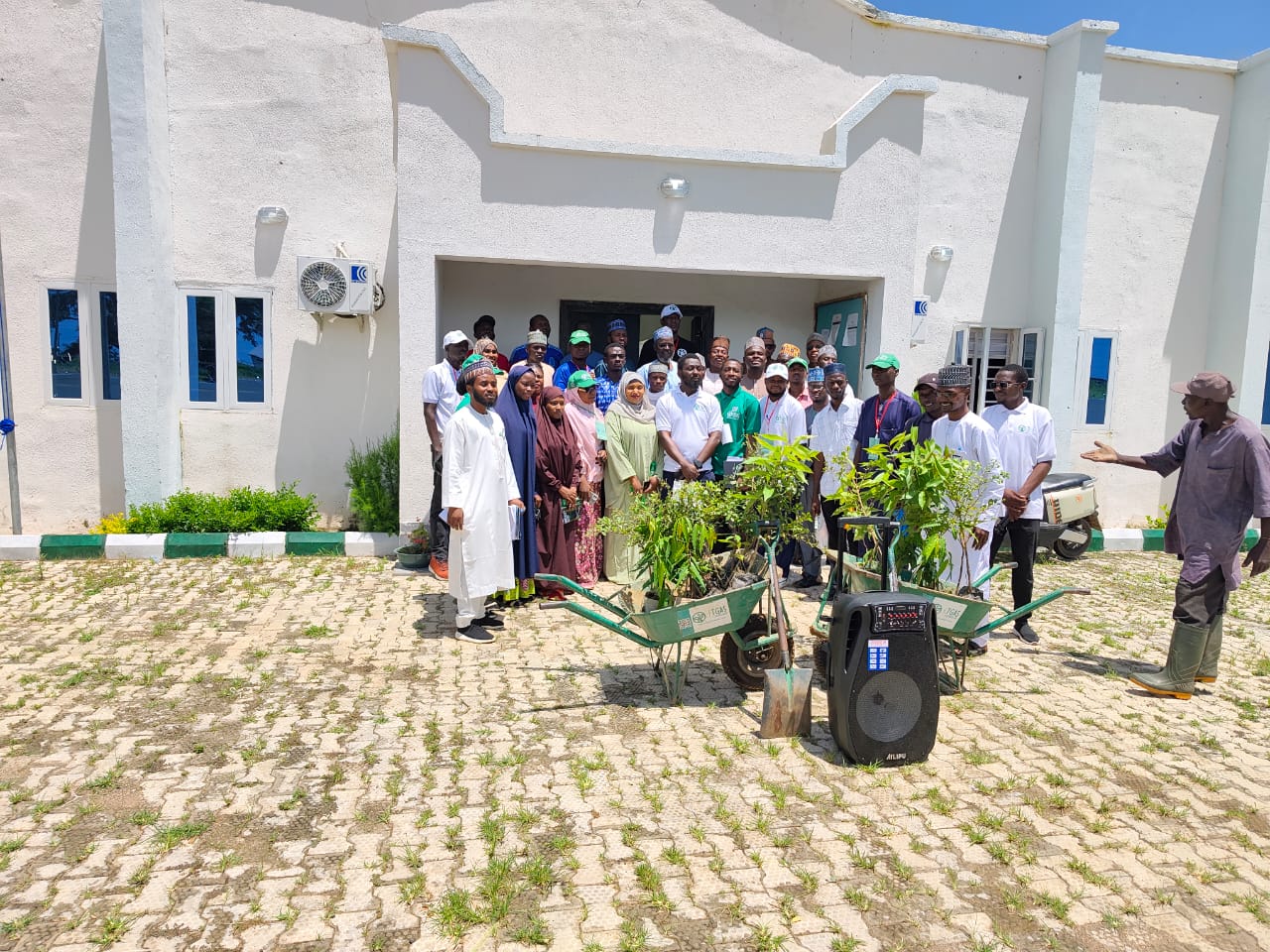 A large group of University staff and students standing in front of a campus building with wheelbarrows of tree seedlings